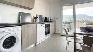 A modern kitchen with a washing machine, dishwasher, and oven. There's a dining area with a table and chairs, and a large window offering a view of the mountains. - Superb T2 Splendid view Ajaccio (AJACCIO)
