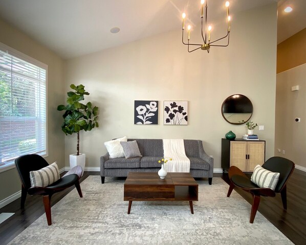 Un salon avec un canapé gris, deux fauteuils, une table basse et un lustre. On y trouve deux tableaux représentant des fleurs, accrochés au mur, une grande plante en pot et un miroir.