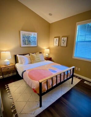 Une chambre à coucher comprenant un lit, deux tables de chevet, une commode, une fenêtre avec des stores et des œuvres d’art murales.