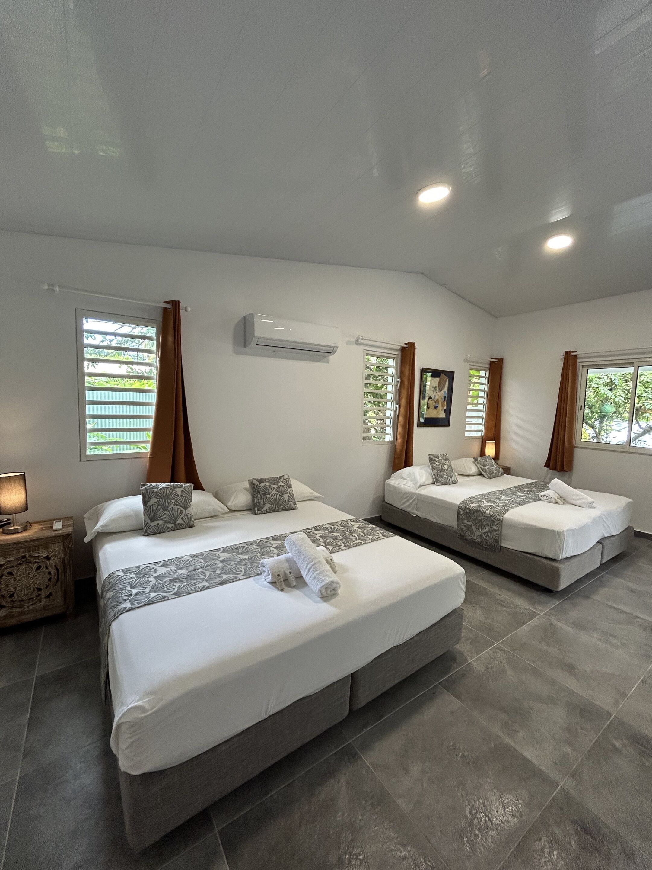 A hotel room with two beds, a window with blinds, an air conditioning unit, and a nightstand with a lamp.