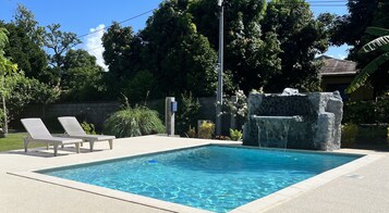 A rectangular swimming pool with a waterfall feature, surrounded by a paved area with two lounge chairs.