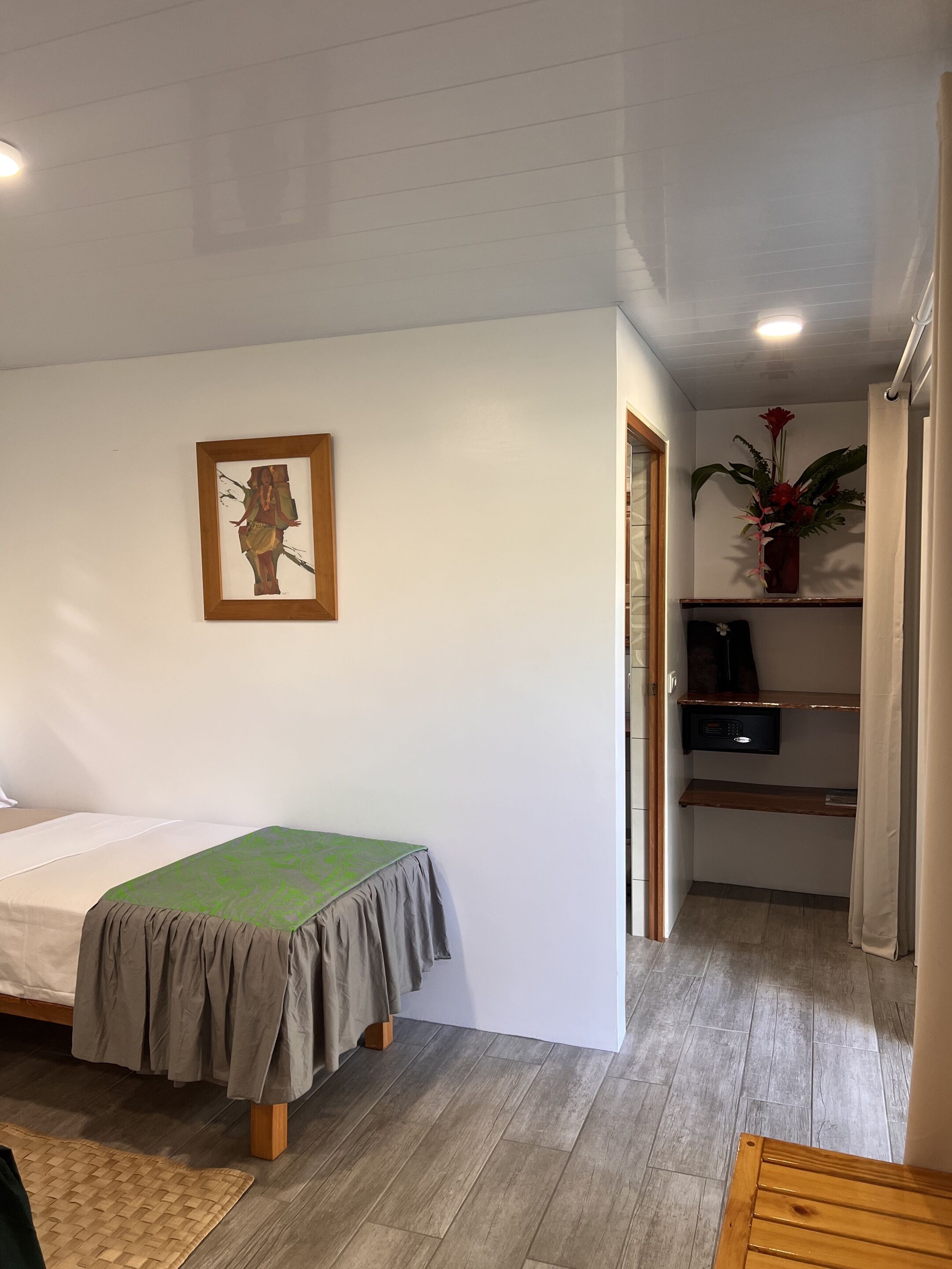 A bedroom with a bed, a small desk, a shelf with a plant, and a painting on the wall.