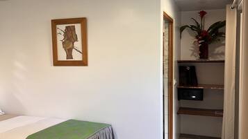 A bedroom with a bed, a small desk, a shelf with a plant, and a painting on the wall.