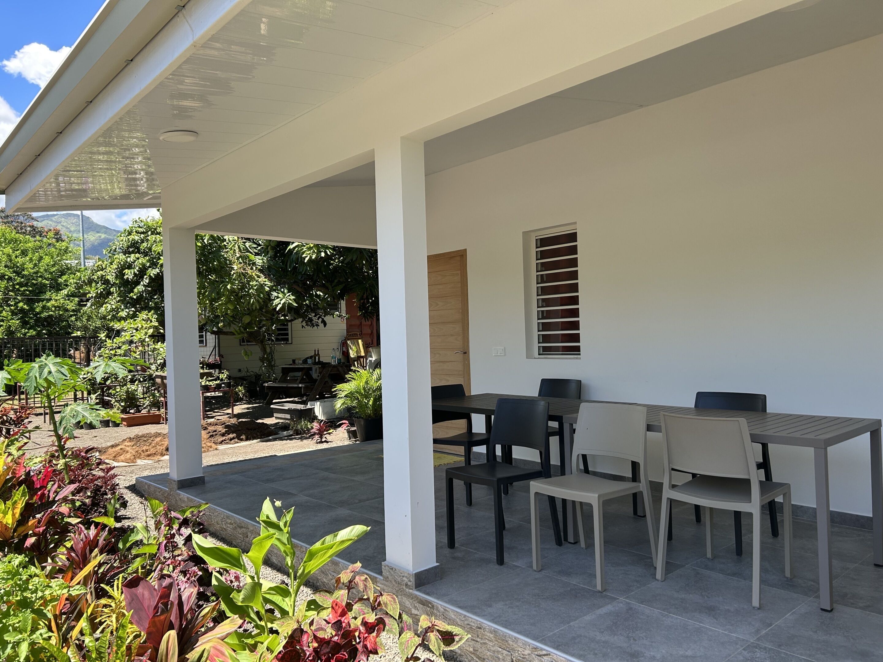 A covered patio area with a table and chairs, surrounded by potted plants and a view of a garden.