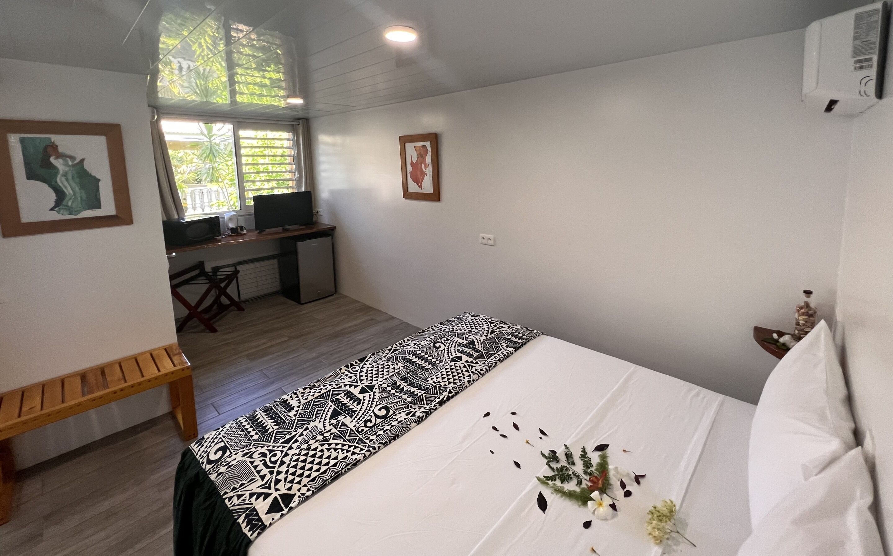 A bedroom with a bed, a desk, and a chair. There is a window with blinds, a picture on the wall, and a small table with a bottle and a plant.