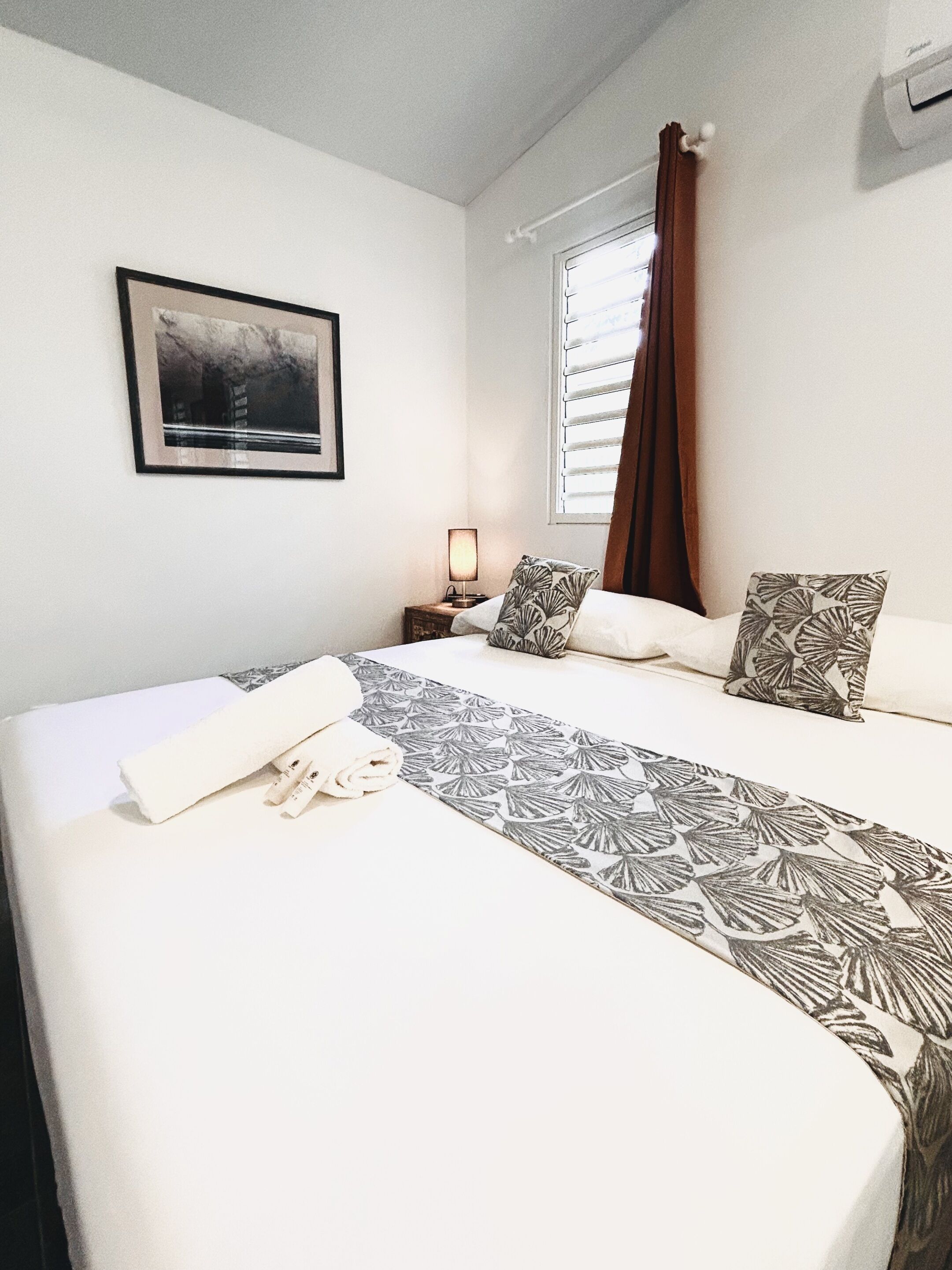 Een net opgemaakt bed met beddengoed met een patroon, een bedlampje, een ingelijst foto aan de muur en een raam met gordijnen.