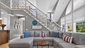 A modern living room with a sectional sofa, a round coffee table, and a staircase with a railing. - Amazing Home Near IU's Campus with INDOOR Basketball Gym & Outdoor Pool/Hot tub (Bloomington)