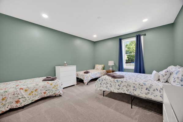 A bedroom with three beds, a white dresser, a window with blue curtains, and a ceiling light.