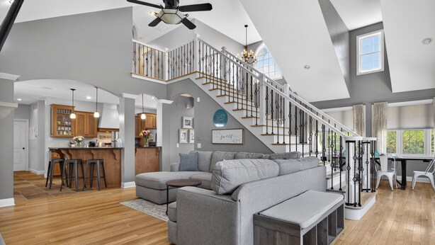 A spacious living area with a gray sectional sofa, a wooden floor, a staircase with a black railing, and a kitchen in the background. - Amazing Home Near IU's Campus with INDOOR Basketball Gym & Outdoor Pool/Hot tub (Bloomington)