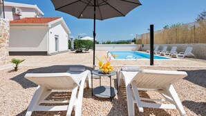 A poolside area with two white lounge chairs, a table with fruit and drinks, and a large umbrella. - Amazing home in Zmijavci (Zmijavci)