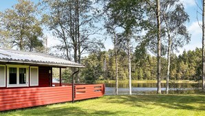 Een rode blokhut met een metalen dak, gelegen op een grasveld met een meer en bomen op de achtergrond.