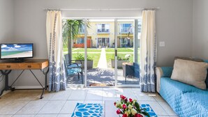 A living room with a TV, a sofa, a coffee table with flowers, and a view of a garden and building through glass doors. - Gulf Bliss: 5MinWalkToBeach,Pool,Yard,CloseToAll (Miramar Beach)