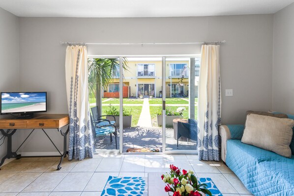 A living room with a TV, a sofa, a coffee table with flowers, and a view of a garden and building through glass doors.