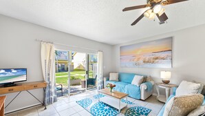 A modern living room with a TV, sofa, coffee table, and a painting of a sunset. - Gulf Bliss: 5MinWalkToBeach,Pool,Yard,CloseToAll (Miramar Beach)