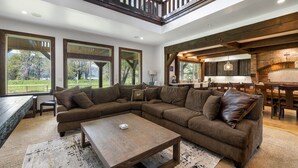 A spacious living room with a large sectional sofa, a coffee table, and a fireplace. There are large windows offering a view of the outdoors.