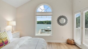 Une chambre avec une grande fenêtre, un lit, une table de chevet, une lampe, un miroir et une vue sur un quai et l’eau.