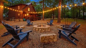 A rustic cabin with a stone fire pit and Adirondack chairs in a wooded area.