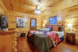 A wooden cabin bedroom with a bed, two lamps, a ceiling fan, and artwork on the walls. - The Arrington,Outdoor Area,Playset,Hot Tub/Fire Pit (Broken Bow)