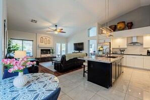 A spacious living area with a sofa, coffee table, and a vase of flowers. There is a kitchen with a large island, cabinets, and a ceiling fan.