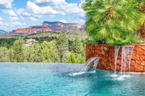 A swimming pool with a waterfall feature, surrounded by lush greenery and a mountainous landscape in the background.