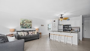 A modern living room with a grey sofa, a kitchen with white cabinets, and a ceiling fan.
