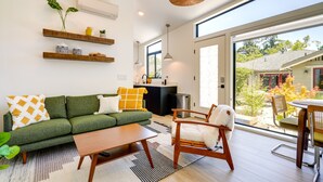 Een moderne woonkamer met een groene bank, een houten salontafel en een rieten fauteuil. Er staan planken met decoratieve spullen en een groot raam met uitzicht op de omgeving.