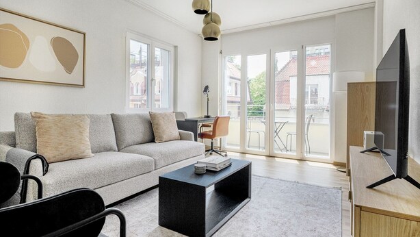 Una sala de estar moderna con sofá, mesa de centro y televisión. Se observan ventanas grandes y un balcón.
