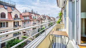 Un balcón con una mesa y sillas de madera, con vistas a una hilera de edificios tradicionales europeos con balcones y tejados de tejas rojas.