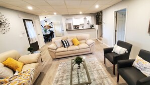 A spacious living room with a beige sofa, a black armchair, and a patterned rug. A dining area is visible in the background with a kitchen. - Updated, Spacious, Fully Fenced Yard (Northfield)