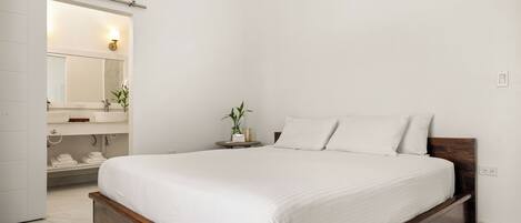Un dormitorio con estructura de cama de madera, colchón blanco, mesita de noche con planta y baño blanco con espejo y toallero.