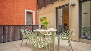 Een klein terras buiten, met een ronde tafel en vier stoelen, een vaas met bloemen en een raam met uitzicht op het interieur.