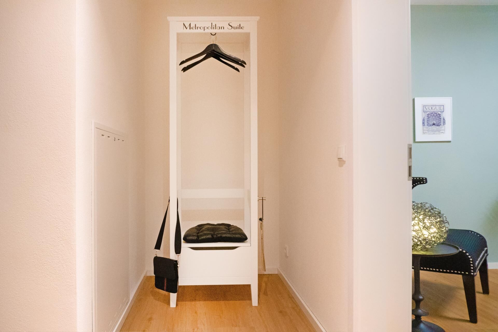 Un couloir étroit avec un placard blanc portant l’inscription « Suite Métropolitaine », contenant une tringle à vêtements et une étagère sur laquelle se trouve un sac noir.