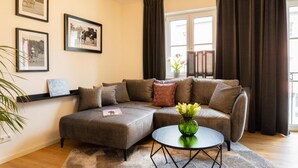 Een moderne woonkamer met een hoekbank, een rond bijzettafeltje en een klein nachtkastje. Er hangen ingelijste foto's aan de muur, staat een potplant en is er een raam met gordijnen.