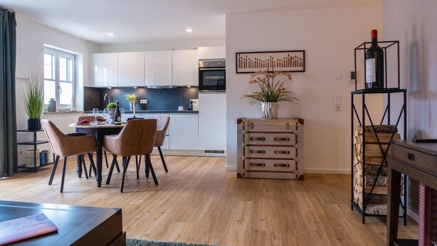 Una cocina moderna con armarios blancos, un comedor con mesa y sillas de madera, y una pequeña mesita con una planta.