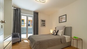 Un dormitorio con cama, una silla, mesita de noche, ventana con cortinas y un muro con cuadros enmarcados.