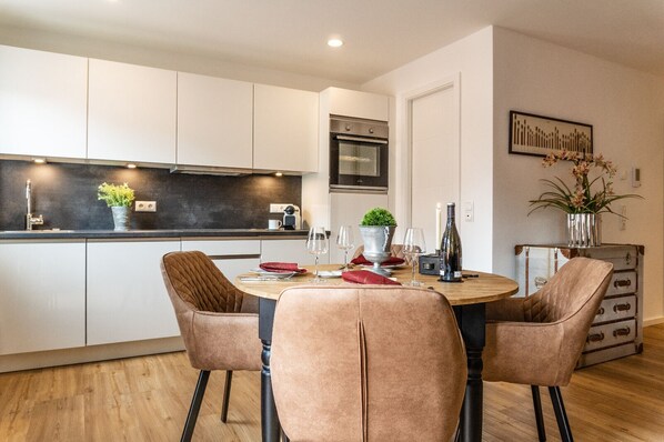 A modern kitchen with white cabinets, a black countertop, and a built-in oven. There is a dining table with four chairs, a vase with flowers, and a wine bottle.