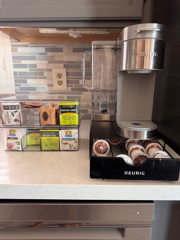 Uma cozinha com cafeteira Keurig, porta-cápsulas de café e diversas cápsulas de chá e café sobre o balcão.