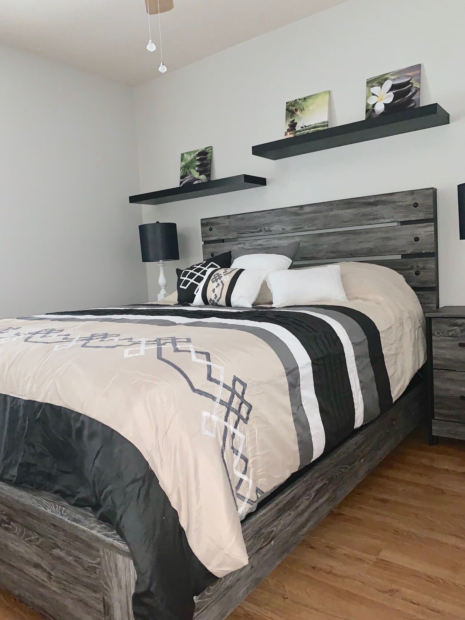 A bedroom with a wooden bed frame, a beige and black patterned bedspread, two bedside tables with lamps, and wall-mounted shelves with decorative items.