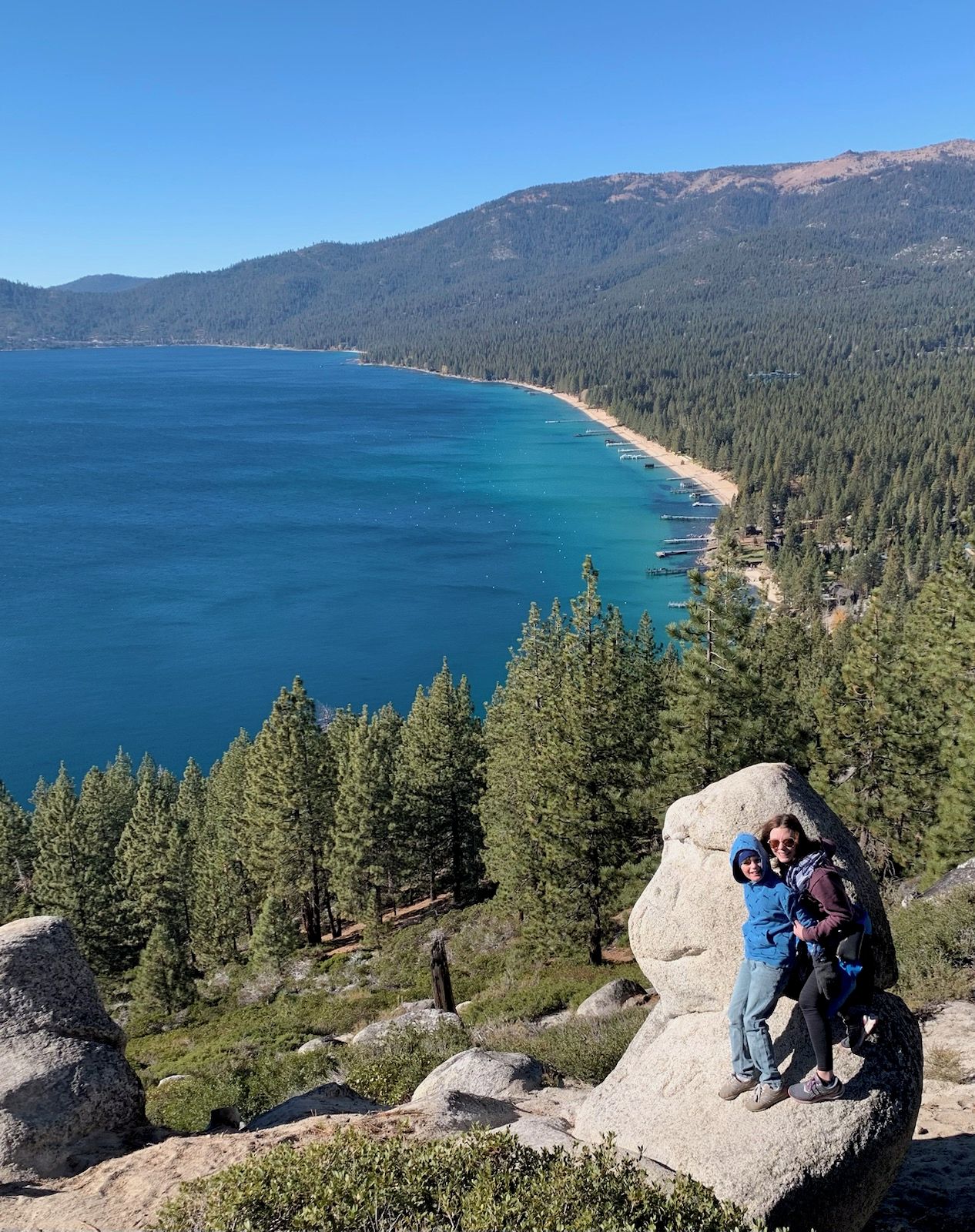 A natural landscape with a clear view of a lake, surrounded by dense forest and mountains.