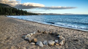 Una playa con un círculo de rocas en la arena, cielo despejado y montañas al fondo.