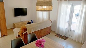 Une pièce comprenant un lit, un téléviseur, une table à manger avec des chaises, un luminaire suspendu en osier et une vue sur l’extérieur par une porte.