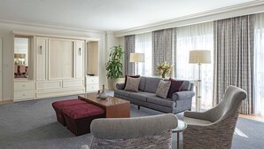 A hotel room with a grey sofa, a red ottoman, a grey armchair, a small side table, a mirror, and a large window with curtains.