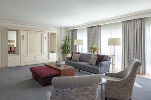 Habitación de hotel con sofá gris, un reposapiés rojo, sillón gris, una mesita auxiliar, un espejo y un ventanal grande con cortinas.
