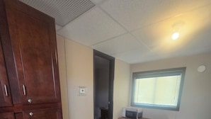 A room with a wooden wardrobe, a desk with a chair, a trash bin, and a window with blinds.