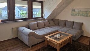 Ein modernes Wohnzimmer mit einer Eckcouch, einem Couchtisch mit Glasplatte und Blick ins Freie durch große Fenster.