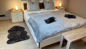 Ein ordentlich bezogenes Bett mit Kissen, eine Nachttischlampe, ein kleiner Holfnachtisch, ein flauschiger Teppich und eine Bank.