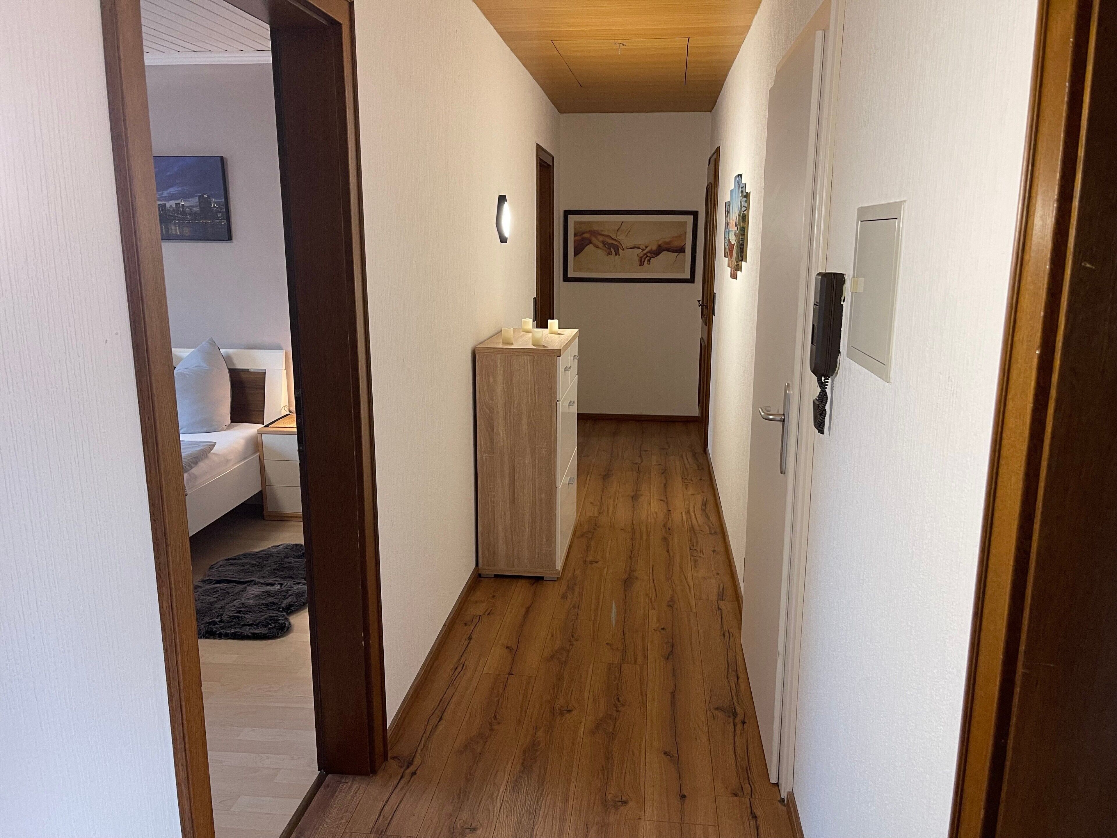 A hallway with wooden flooring, a white wall, a wooden cabinet, and a framed artwork on the wall.