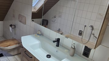 Un bany amb un penjador de fusta, un lavabo blanc, un mirall, un anell per a tovalloles i una dutxa.