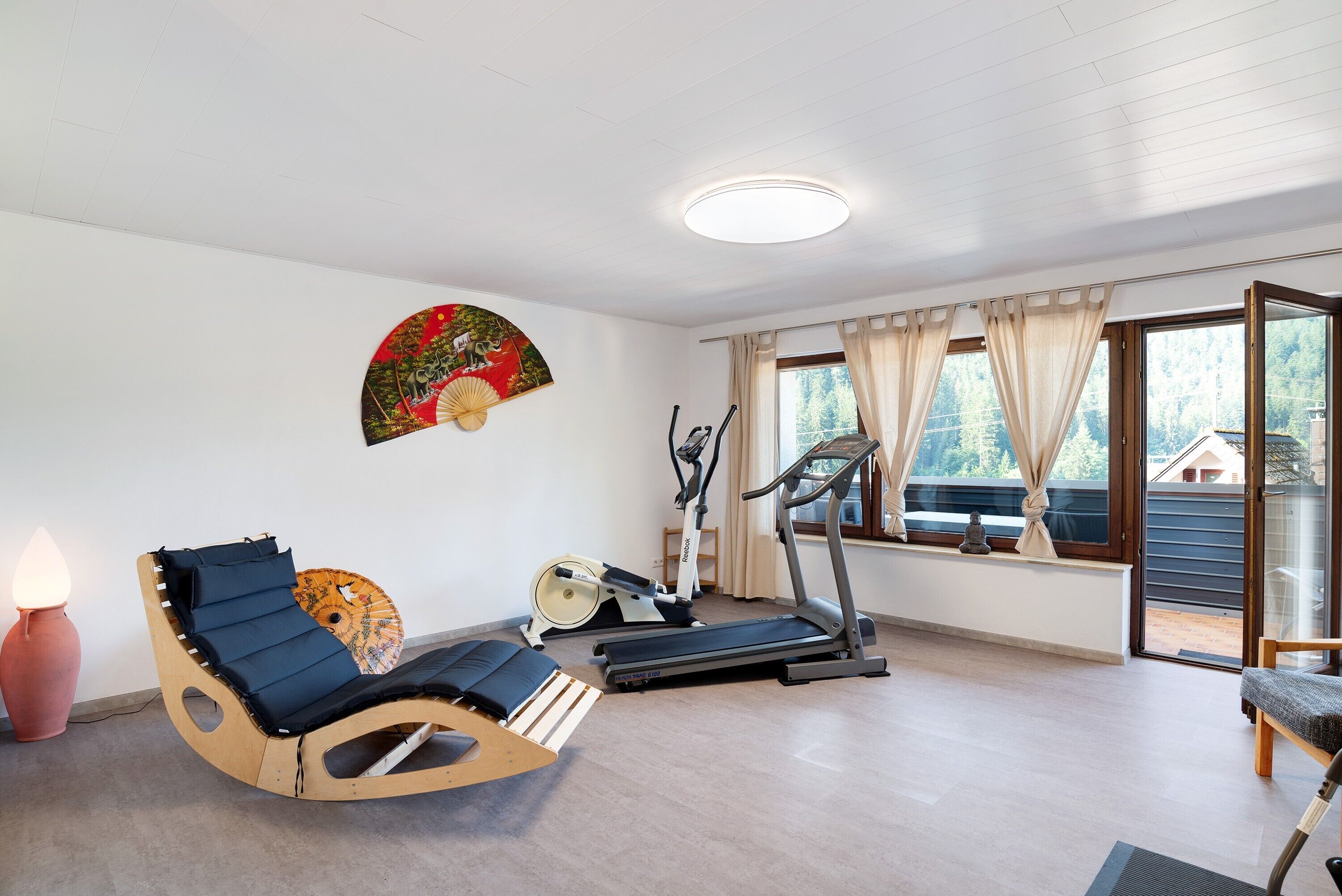 Ein Zimmer mit einem hölzernen Schaukelstuhl, einem Laufband und einem Ventilator an einem Fensterbrett.