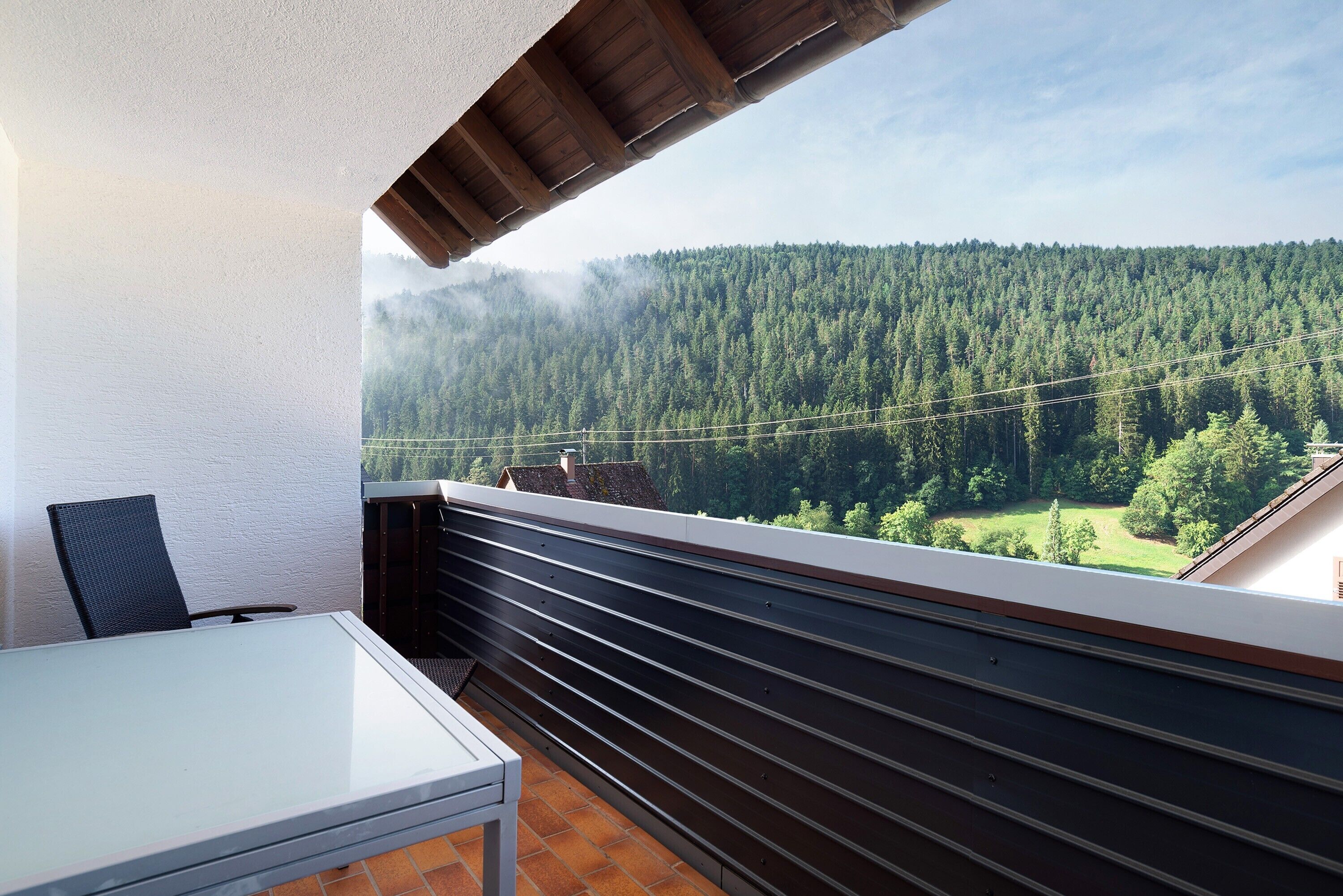 Un balcone con tavolo e sedie, vista su una valle boscosa e una catena montuosa.